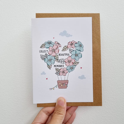 Greeting Card held by hand illustrated with hot air balloon embellished with flowers and has lettering "Collect Beautiful Memories"
