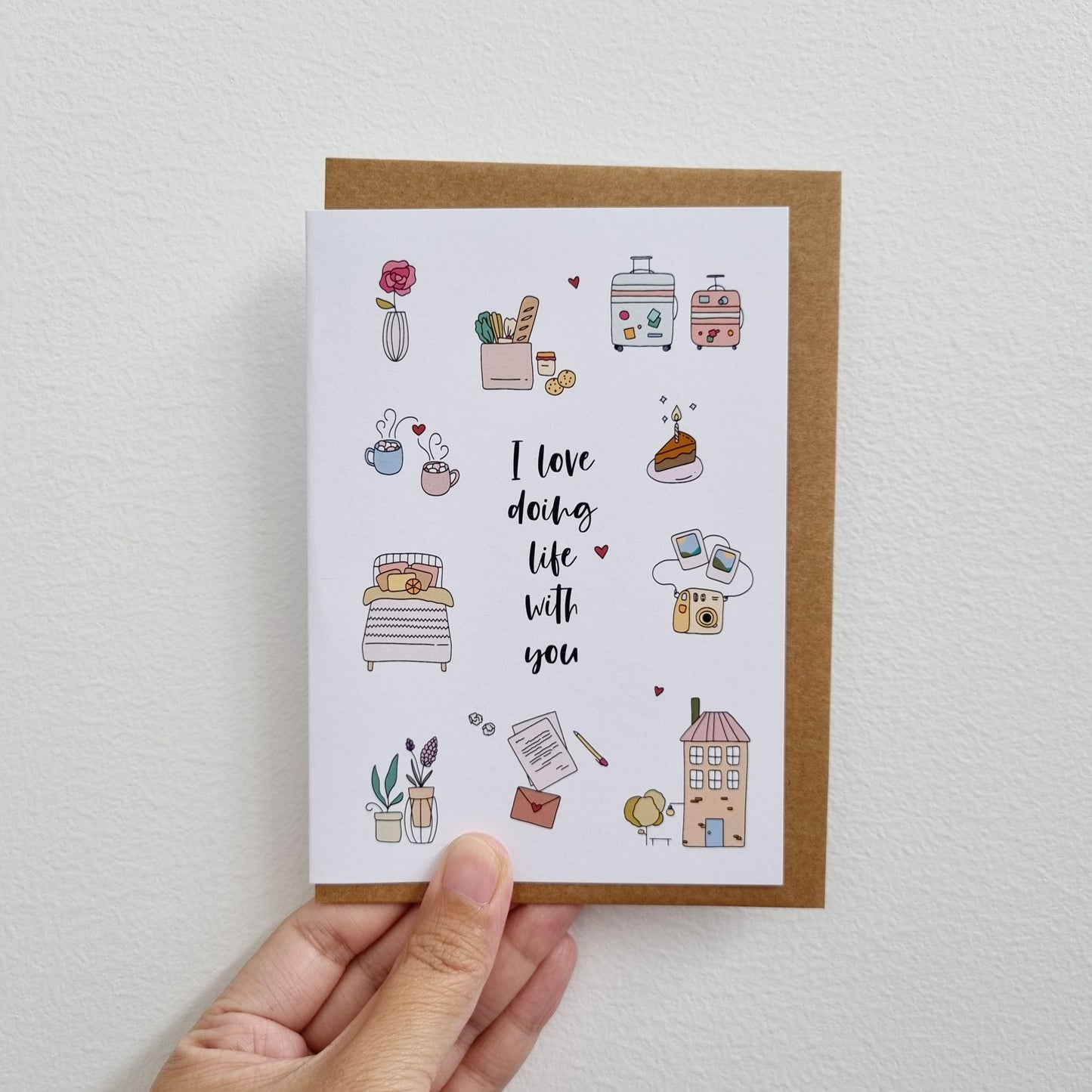 Greeting card with illustration being held by a hand. Illustration has words in the middle "I love doing life with you" surrounded by illustration of flower in vase, groceries, two cups with marshmallow, cosy bed, love letter with pen, house, polaroid camera with two photos, slice of birthday cake, two luggage trolley bags.
