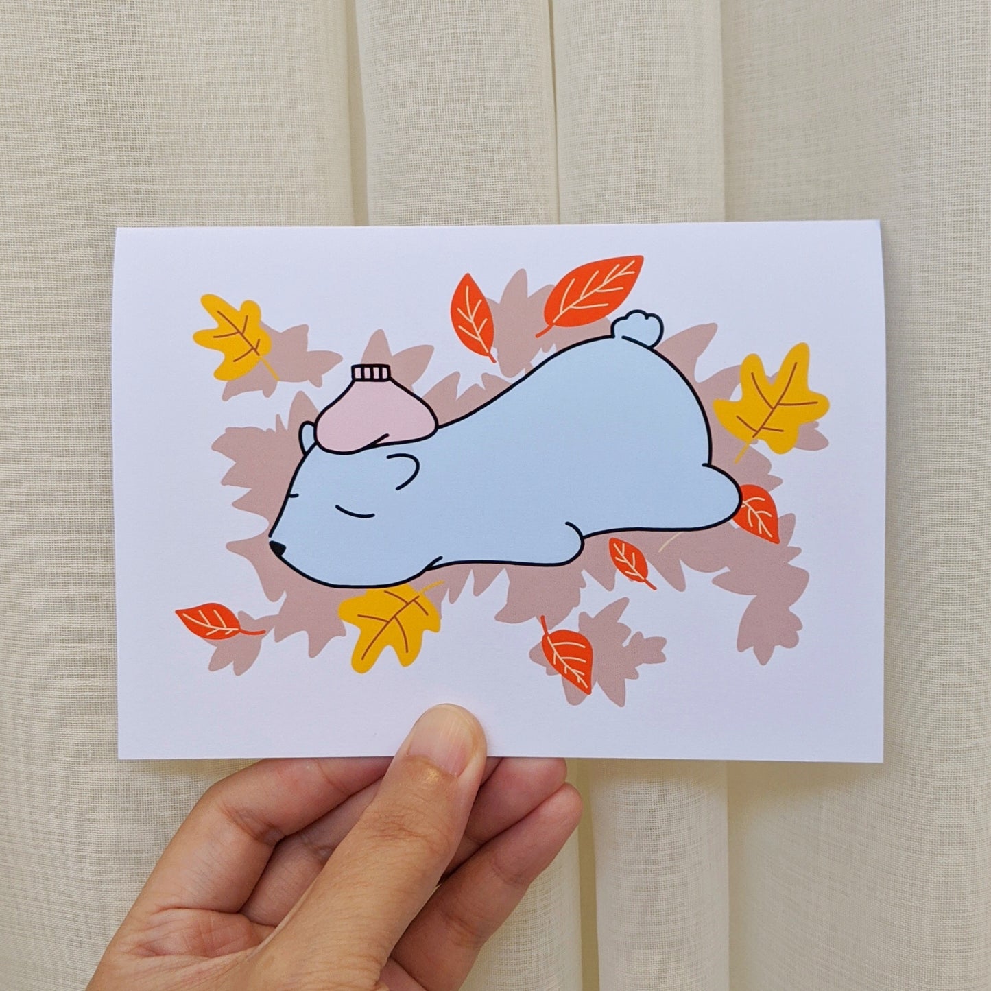 Greeting card held by hand with illustration of polar bear lying tummy down with ice pack over it's forehead surrounded by autumn leaves