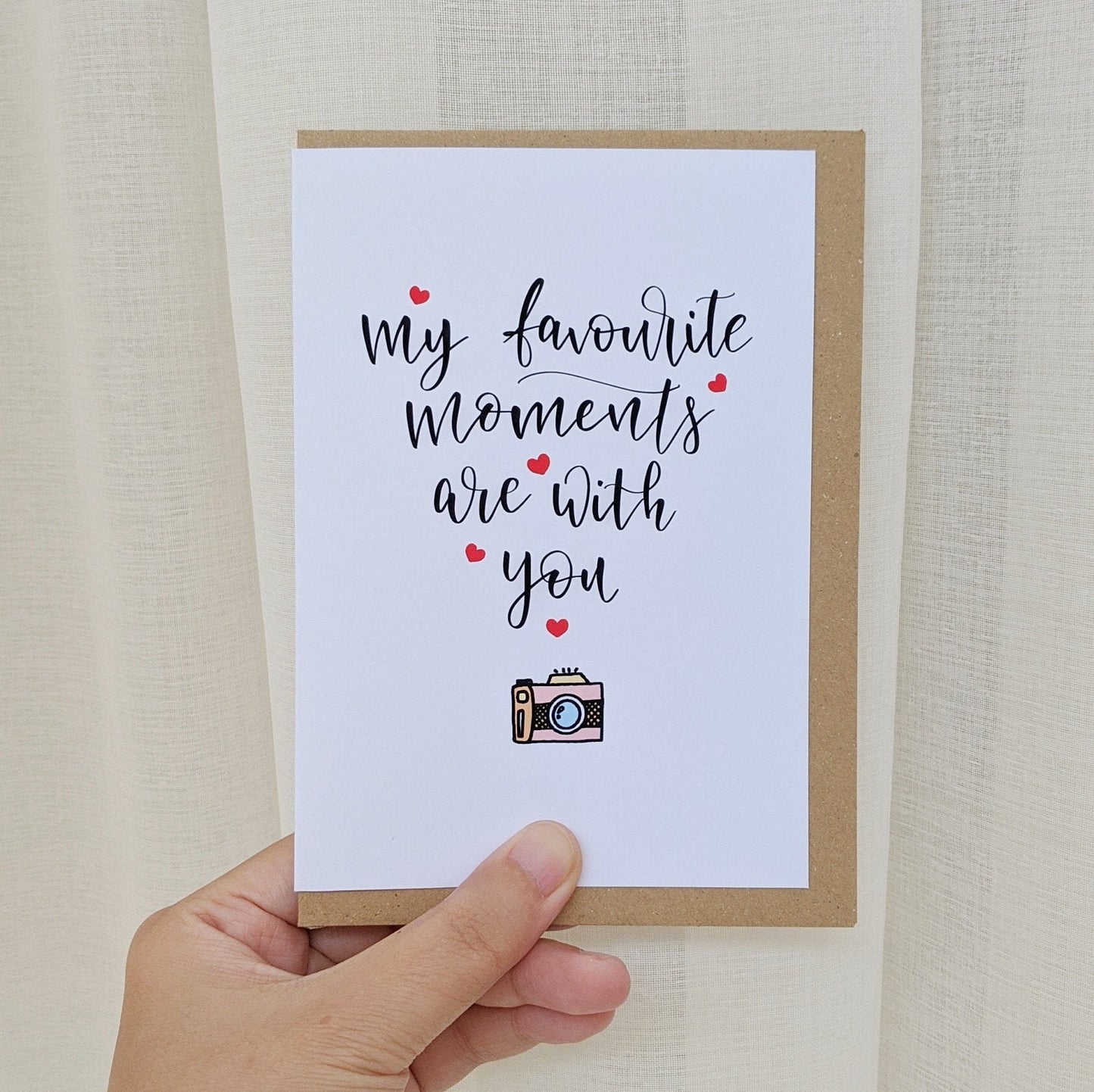 Greeting card held by hand with lettering "my favourite moments are with you" surrounded by a small scatter of love hearts and illustration of camera at the bottom