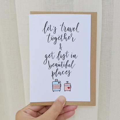 Greeting card being held by hand with lettering "let's travel together & get lost in beautiful places" with illustration of two luggage bags, one in pink and one in blue