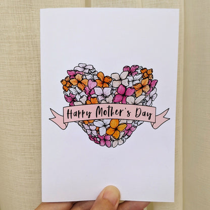 Greeting card held with hand with illustration of a heart flower bouquet with a banner across it with lettering "Happy Mother's Day"