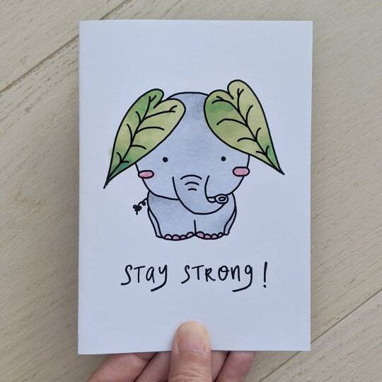 Greeting card held by hand with illustration of elephant embellished with leaf ears and lettering "stay strong" at the bottom