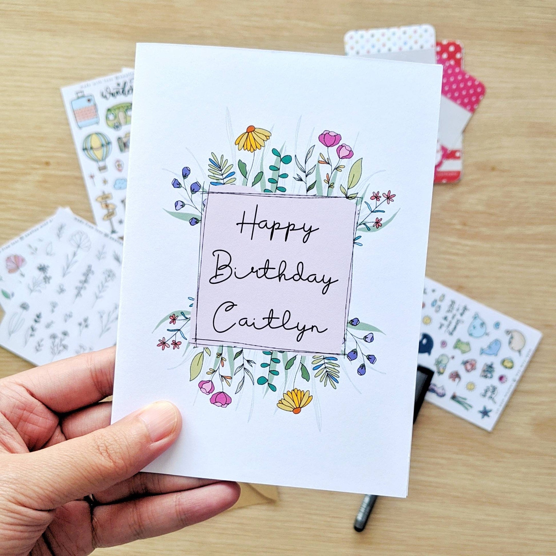Greeting card held by the hand with illustration of a  with pink square surrounded by colourful flowers. Middle of square has the words "Happy Birthday Caitlyn"