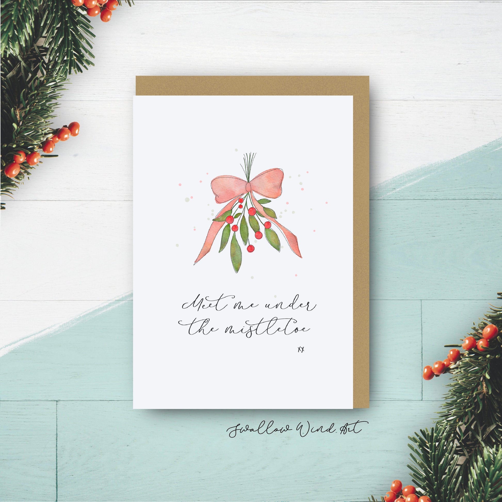 Greeting card with lettering "Meet me under the mistletoe xx" with watercolour illustration of mistletoe tied with red ribbon