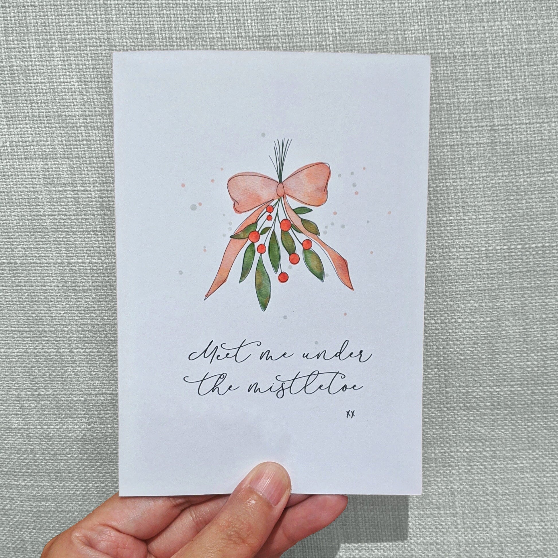 Greeting card held with hand with lettering "Meet me under the mistletoe xx" with watercolour illustration of mistletoe tied with red ribbon
