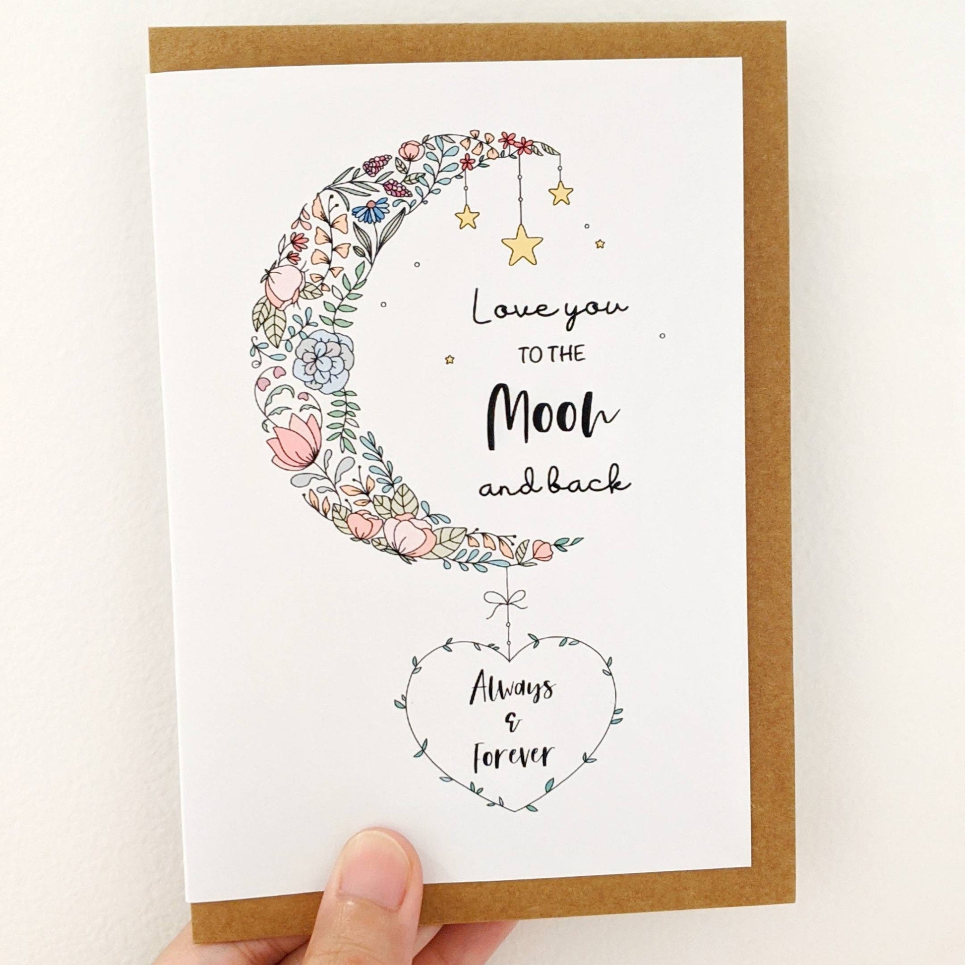 Greeting card held by hand with lettering with words "Love You To The Moon and back" with illustration of moon embelished with flowers and leaves decorated with stars. At the bottom is a leaf heart wreath with wording "Your Text Here"