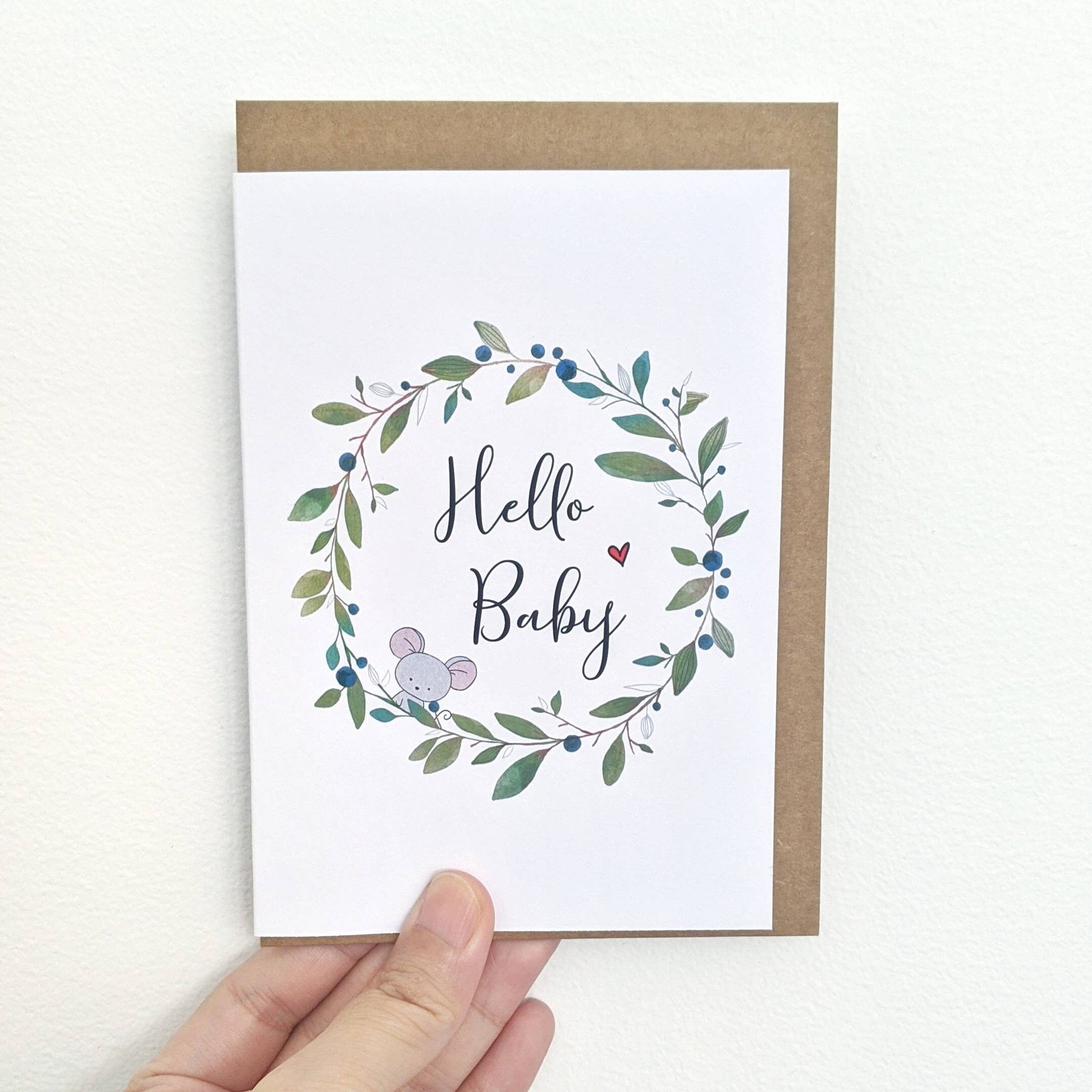 Greeting card with illustration of a wreath being held by a hand. Center of the wreath are the words "Hello Baby" with a little mouse peeping out and red love heart on either side